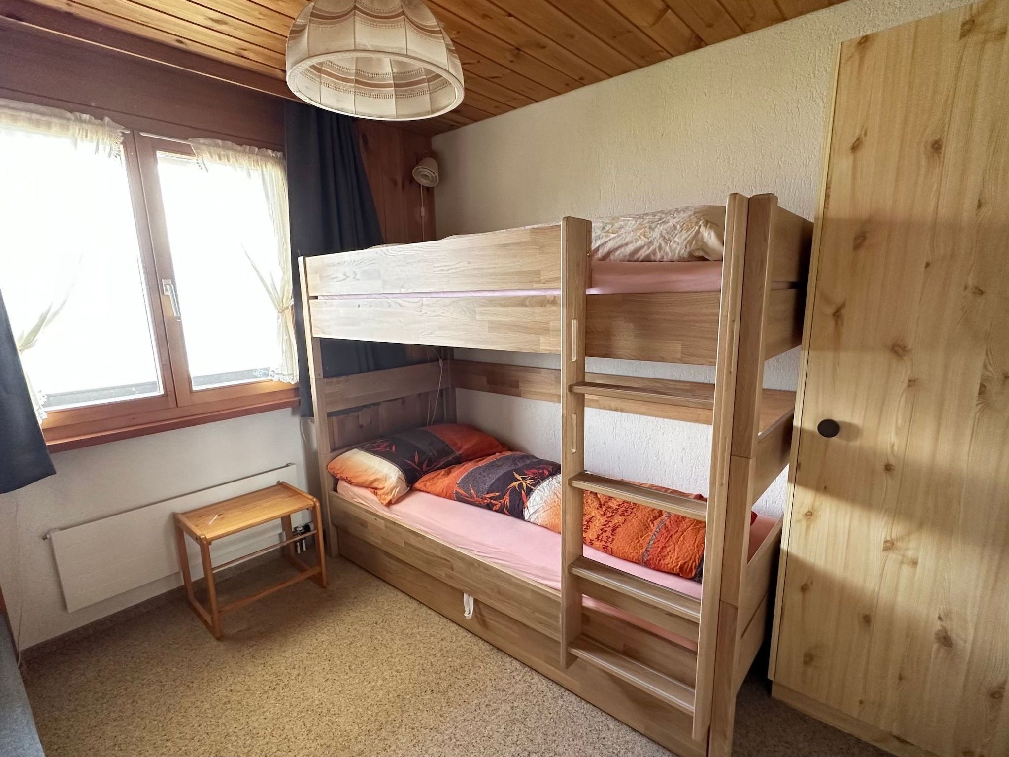 Ein Schlafraum mit Etagenbetten, Holzboden, einem kleinen Tisch und einem Fenster mit Vorhängen.