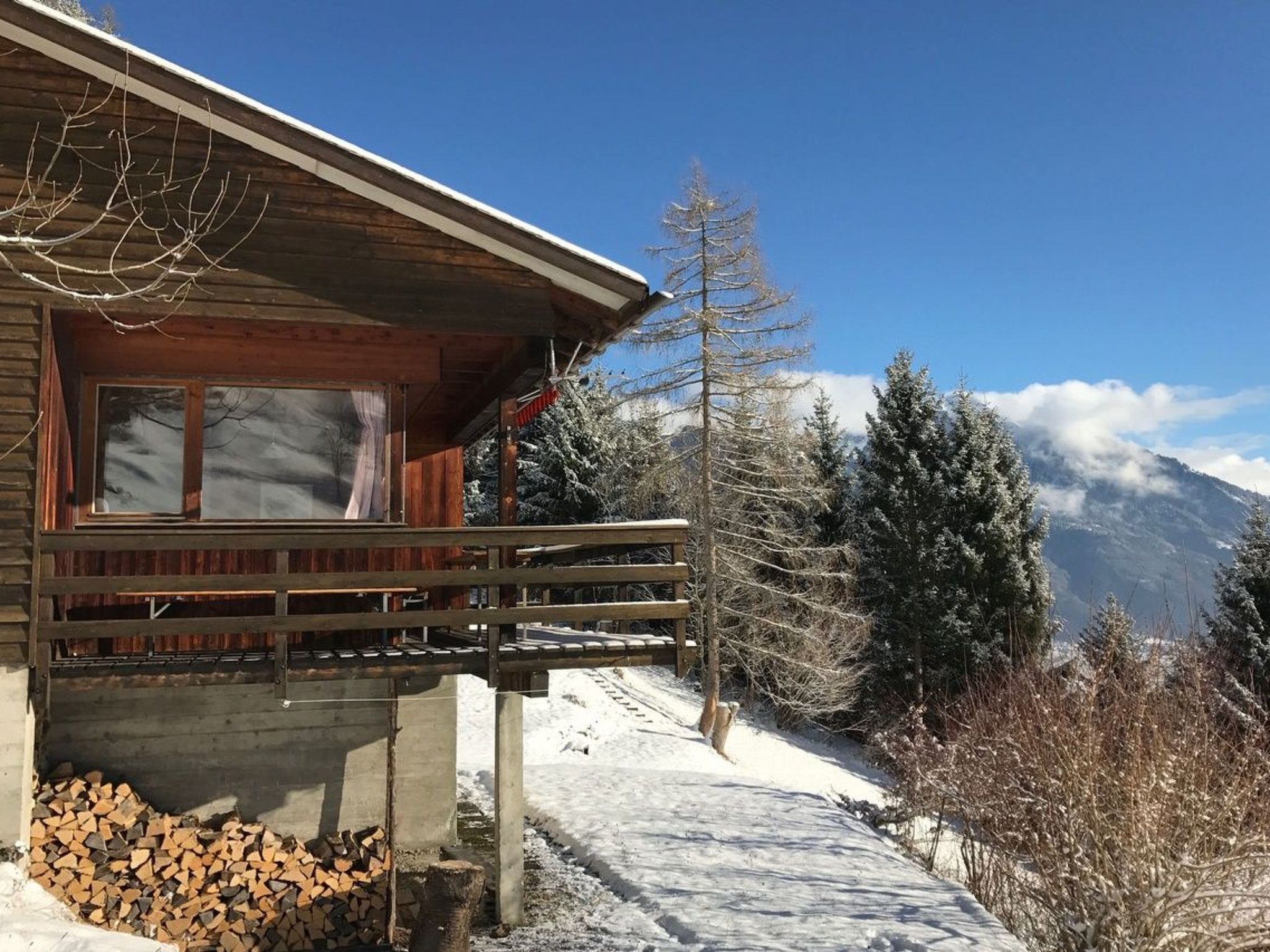 Una cabana de fusta amb un balcó, una finestra i vistes a arbres i muntanyes cobertes de neu.