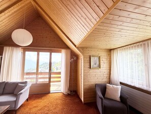 Una habitación acogedora con paredes de madera, un techo con estructura de vigas de madera, un sofá, una silla y un ventanal con cortinas.