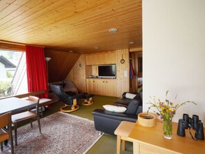A cozy room with a wooden ceiling, a television, a sofa, a coffee table, and a dining area with chairs and a rug.