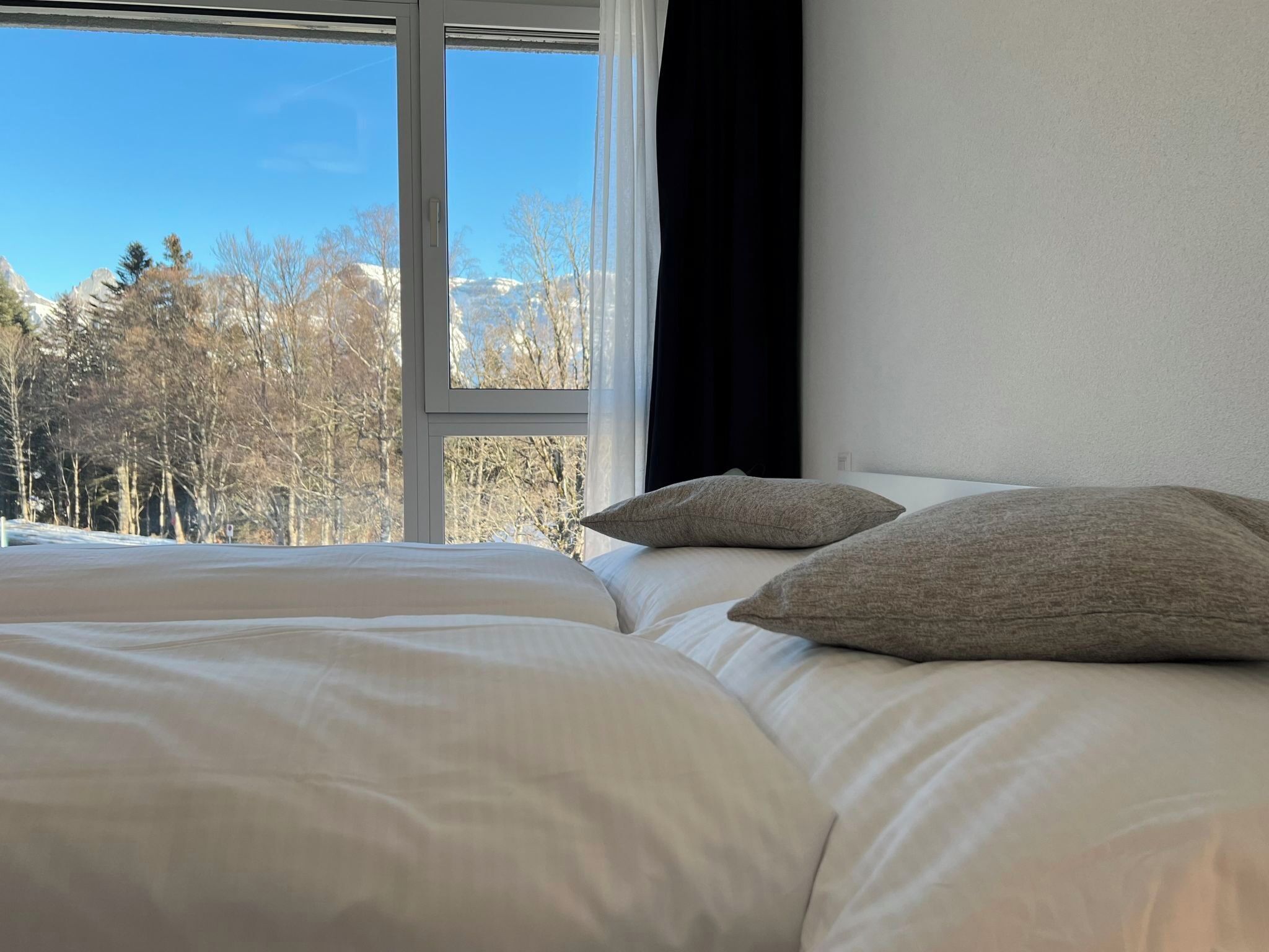 Un lit avec une literie blanche et deux oreillers, une fenêtre donnant sur des arbres et des montagnes, et un rideau noir à la fenêtre.