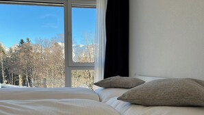 Een bed met witte lakens en twee kussens, een raam met uitzicht op bomen en bergen, en een zwart gordijn voor het raam.
