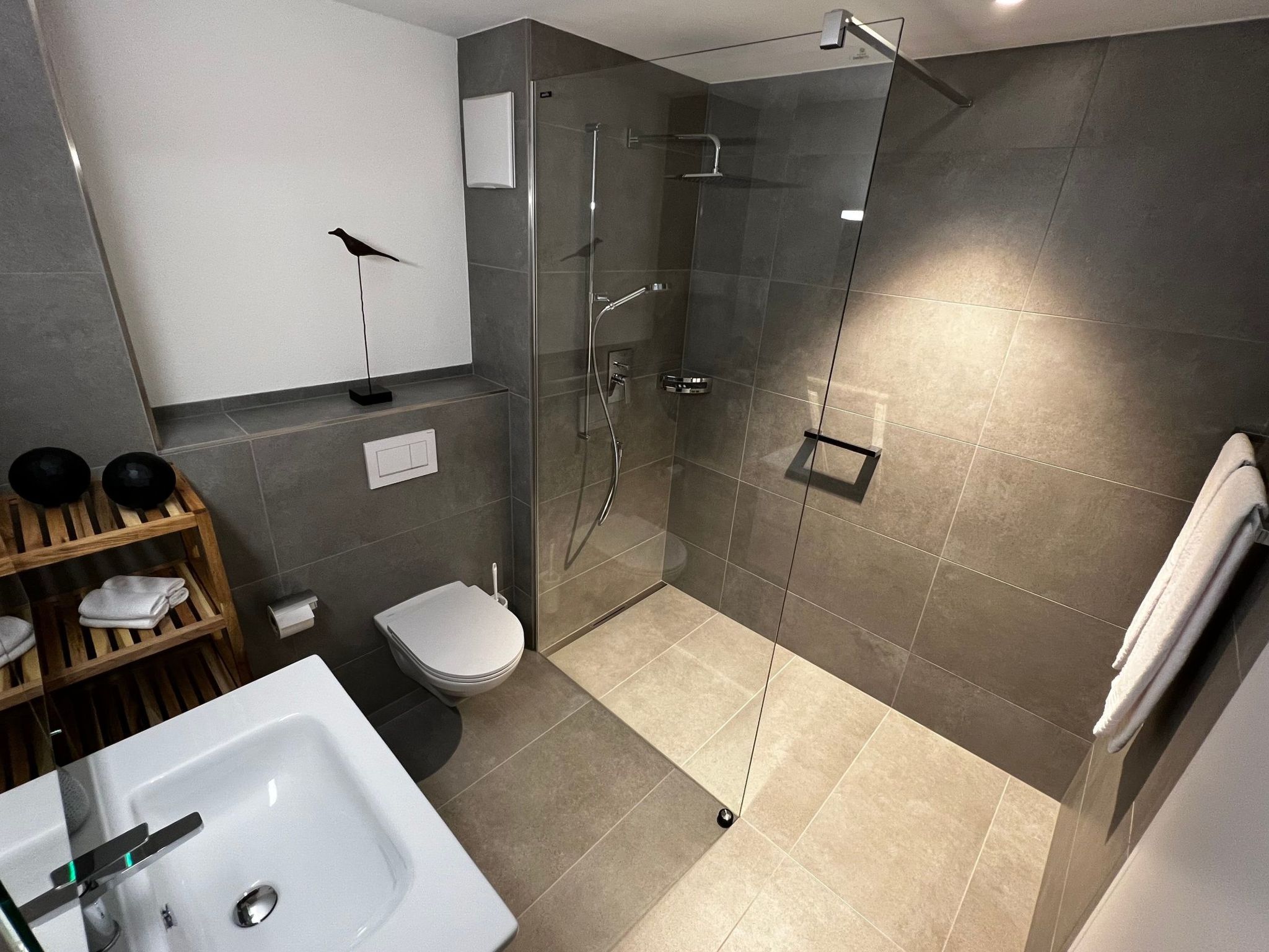 A modern bathroom with a glass-enclosed shower, a toilet, a sink, and a wooden shelf with towels.