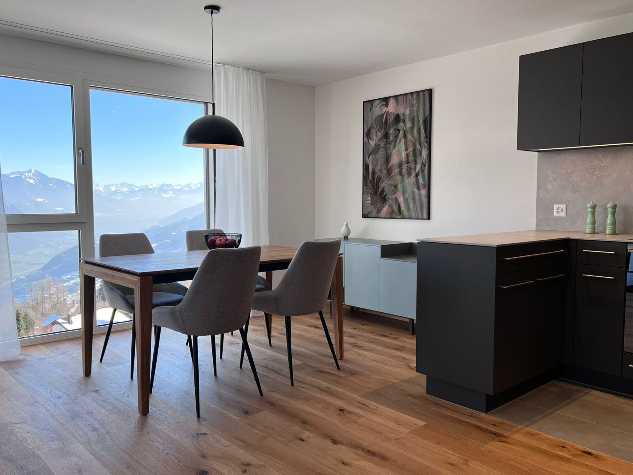 A modern kitchen with a dining area, large windows with a mountain view, and a contemporary art piece on the wall.
