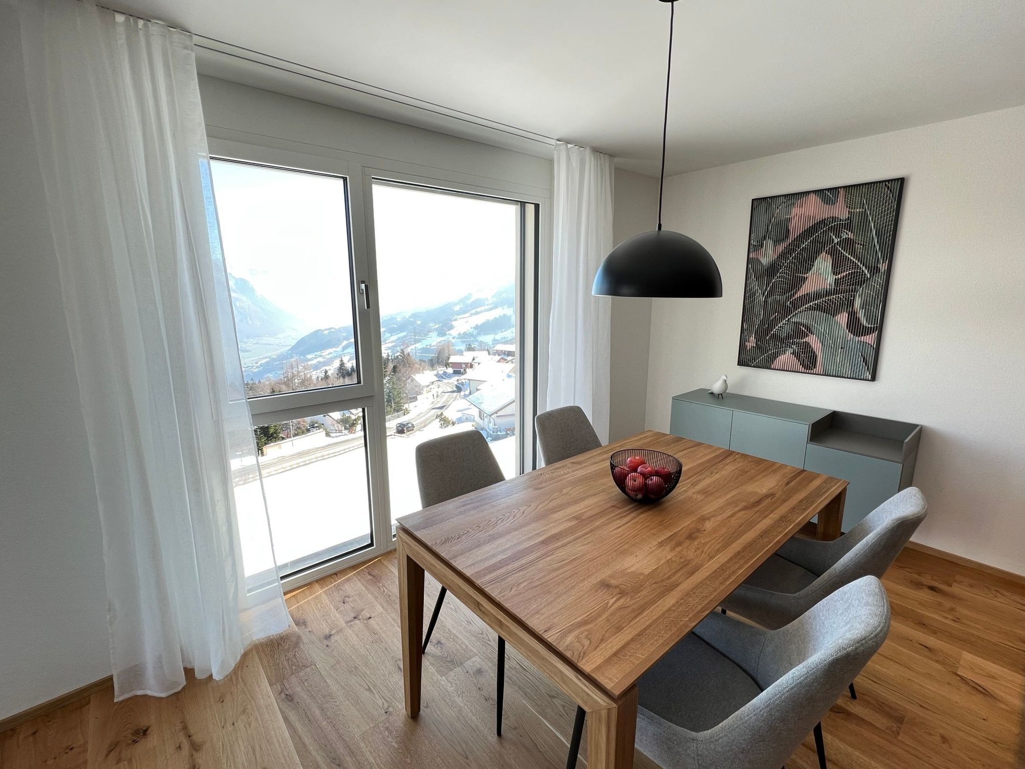 A modern dining area with a wooden table, grey chairs, a black pendant light, and a city view through large windows.