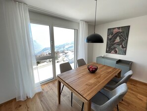 A modern dining area with a wooden table, grey chairs, a black pendant light, and a city view through large windows.
