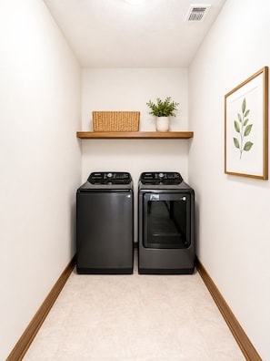 Une buanderie équipée de deux lave-linge et deux sèche-linge à chargement frontal, d’une étagère avec un panier et une plante, et d’une œuvre d’art représentant une feuille, encadrée et accrochée au mur.