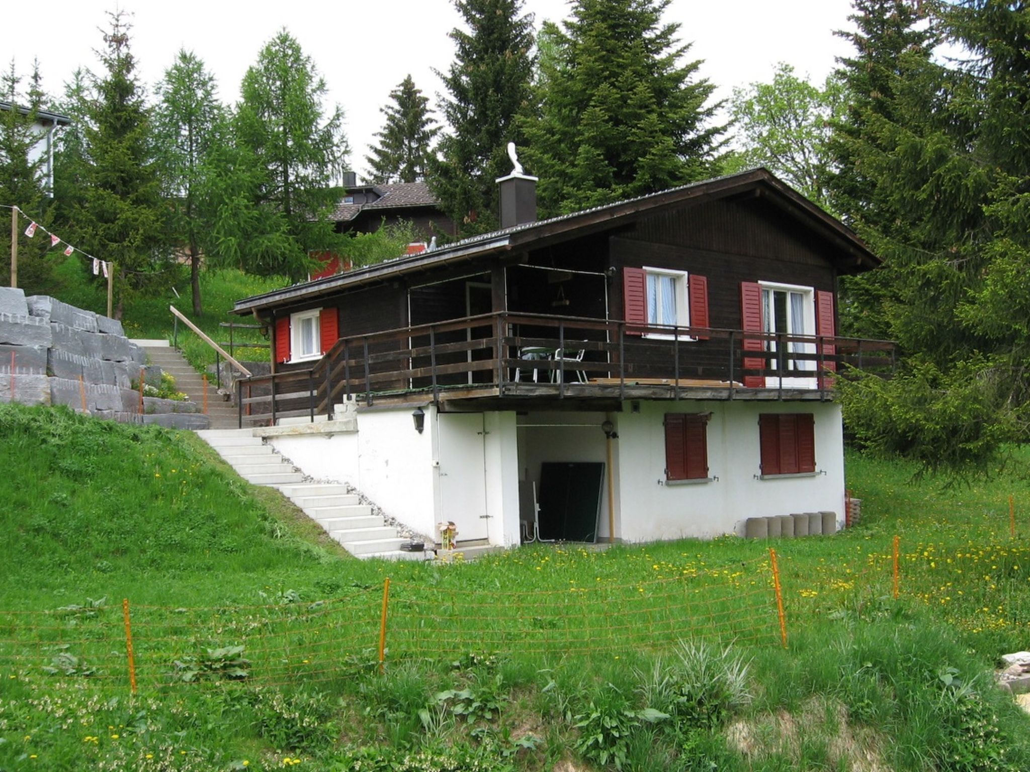A two-story chalet with a balcony, surrounded by greenery and a stone staircase.