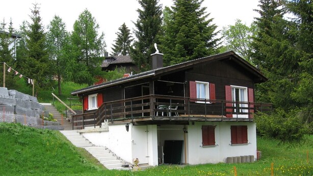 En tvåvånings chalet med en balkong, omgiven av grönska och en trappa av sten.