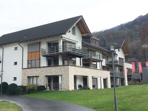 Un edificio de varios pisos con balcones, fachada de piedra y montañas al fondo.