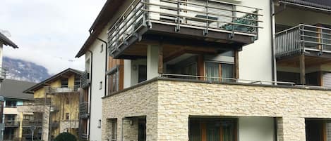 A modern two-story building with a stone facade, balconies, and a view of the mountains.