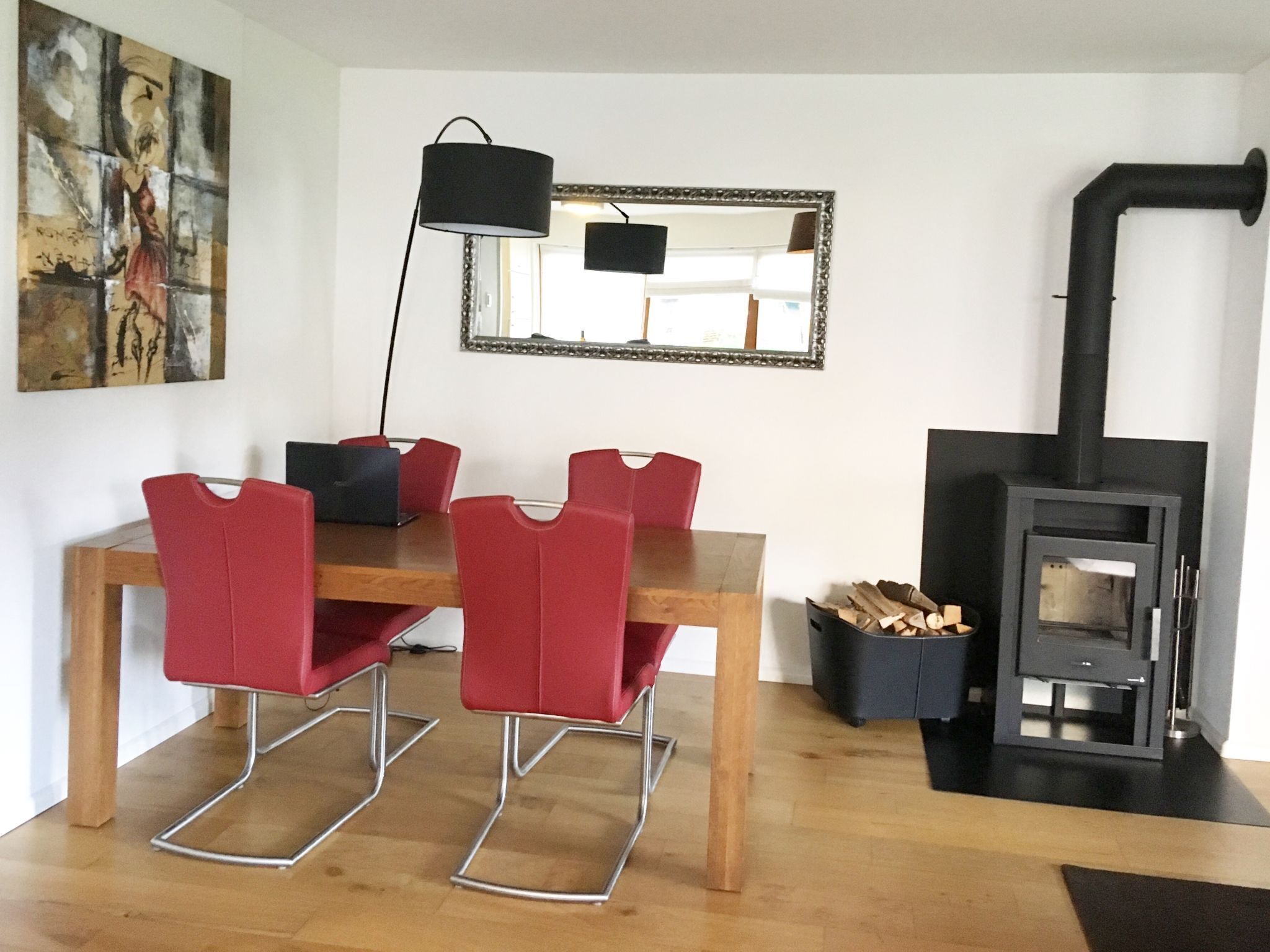 Un coin repas avec une table en bois et des chaises rouges, une lampe noire, un miroir et un poêle à bois.