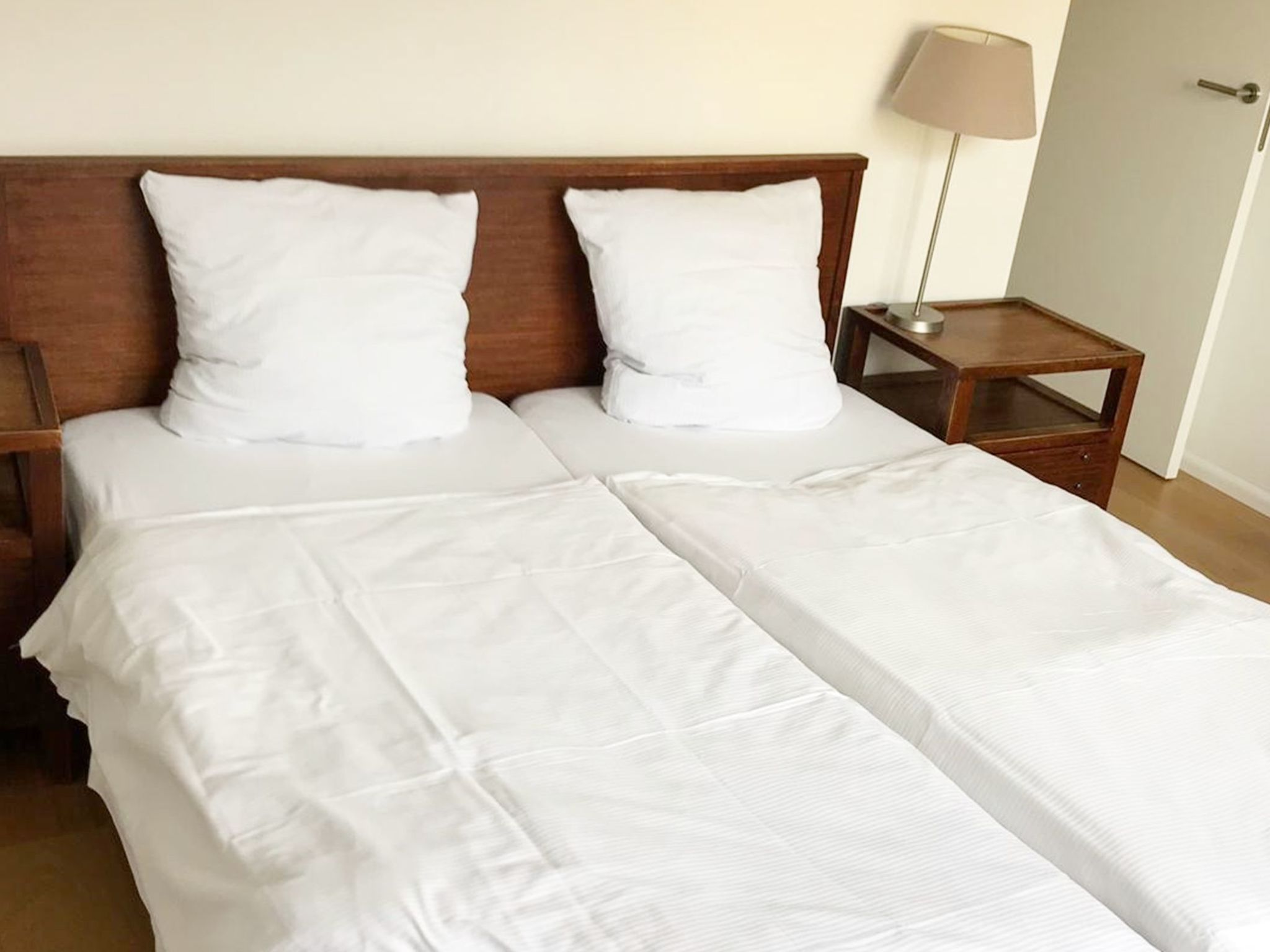 Un lit double avec du linge de lit blanc, une tête de lit en bois, une table de chevet avec une lampe et une commode.