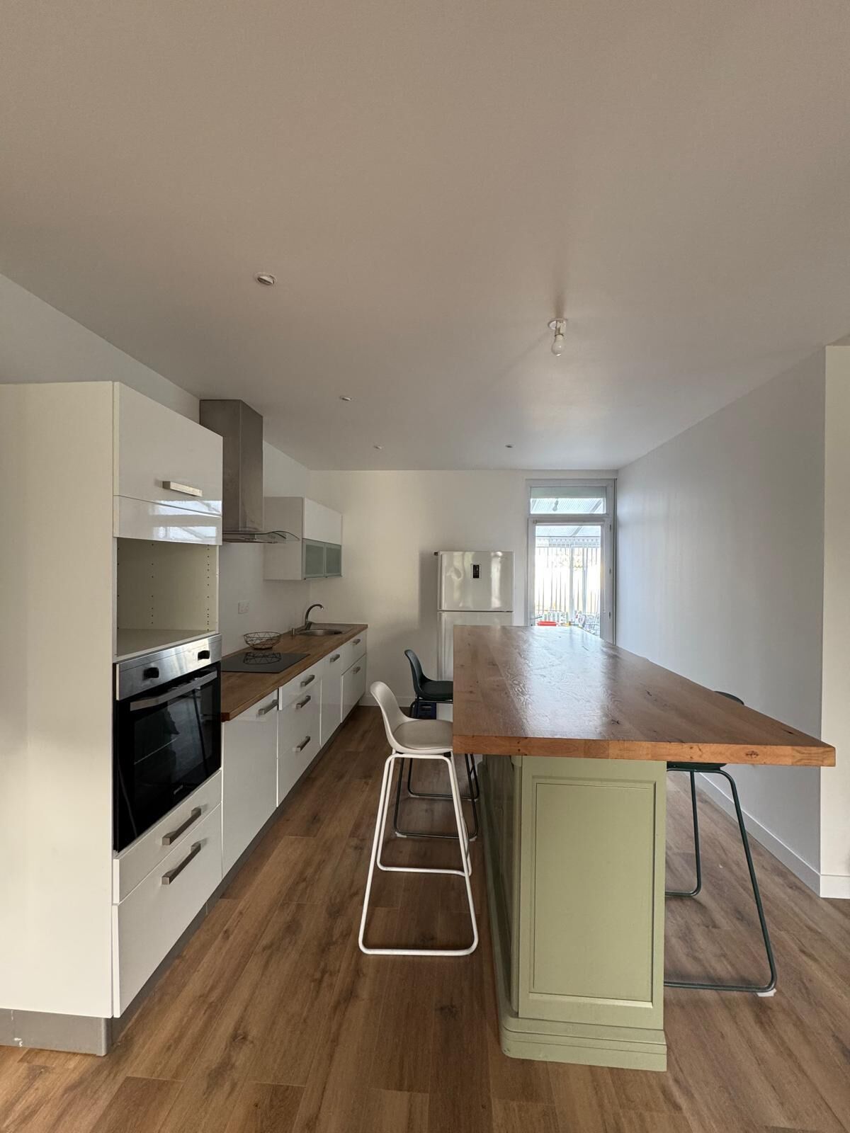 Une cuisine moderne avec des armoires blanches, un plan de travail en bois, un four encastré et un petit îlot central agrémenté d’un tabouret.