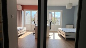 Ett rum med en säng, ett skrivbord och en stol, med utsikt över byggnader utanför fönstret.
