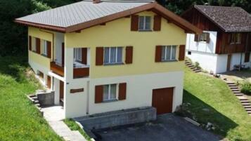 Une maison à deux étages, dotée d’un toit plat, de volets en bois et d’un petit porche à l’avant.