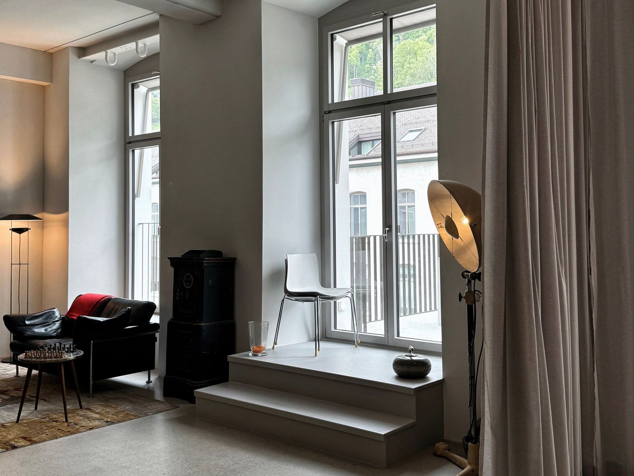 A modern living room with a large window, a black cabinet, a white chair, and a floor lamp.
