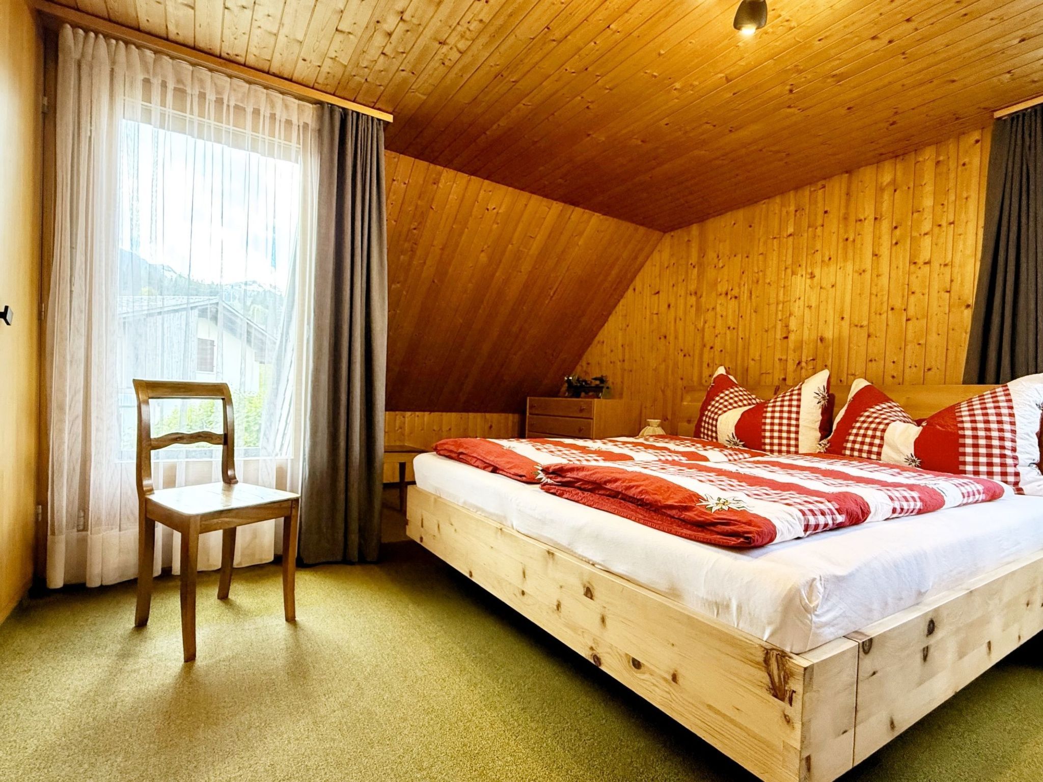 A cozy bedroom with a wooden ceiling, a bed with red and white bedding, a chair, and a nightstand.
