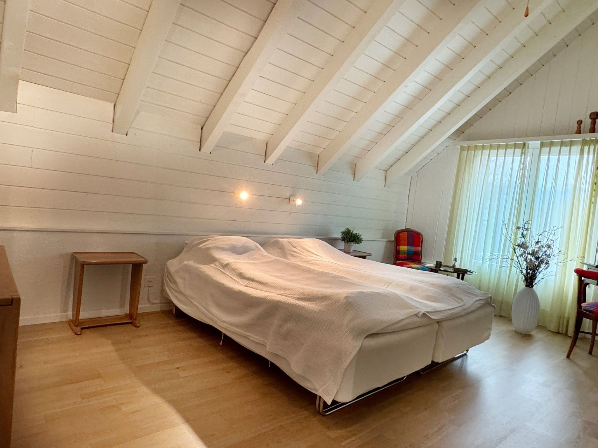 Ein Schlafzimmer mit einem Bett, das mit einem weißen Laken bezogen ist, einem Nachttisch aus Holz, einer kleinen Pflanze und einem Fenster mit Vorhängen.