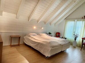 A bedroom with a bed covered in a white sheet, a wooden bedside table, a small plant, and a window with curtains.