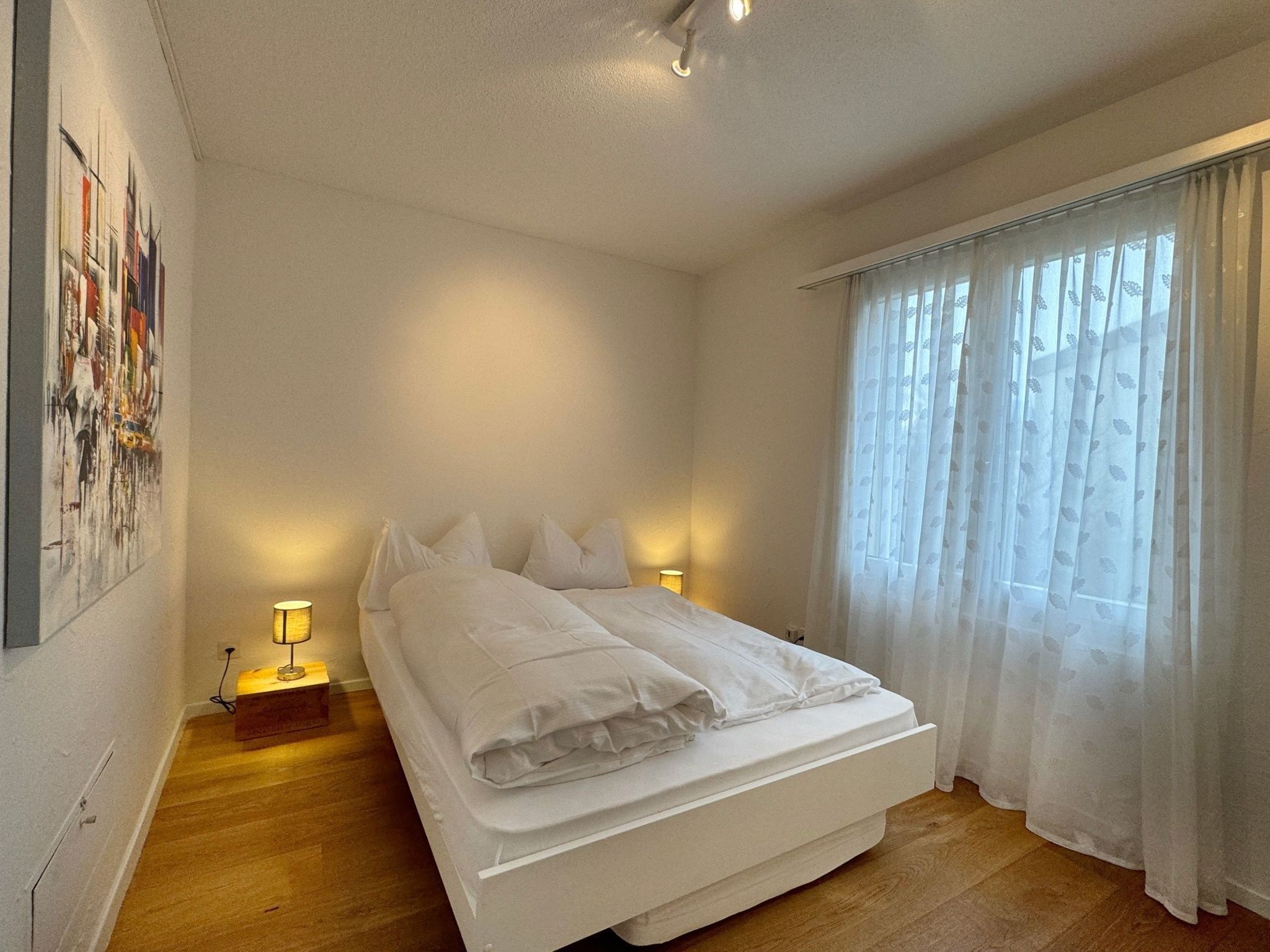 Un dormitorio con cama, lámparas de noche, una ventana con cortinas translúcidas y un cuadro colgado en la pared.