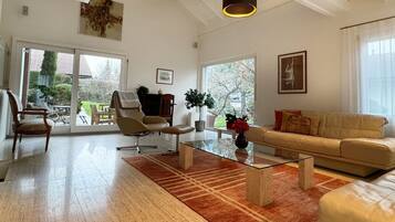 A spacious living room with a glass coffee table, a beige sofa, and a chair. There are two pendant lights, a large window, and a door leading to a balcony.