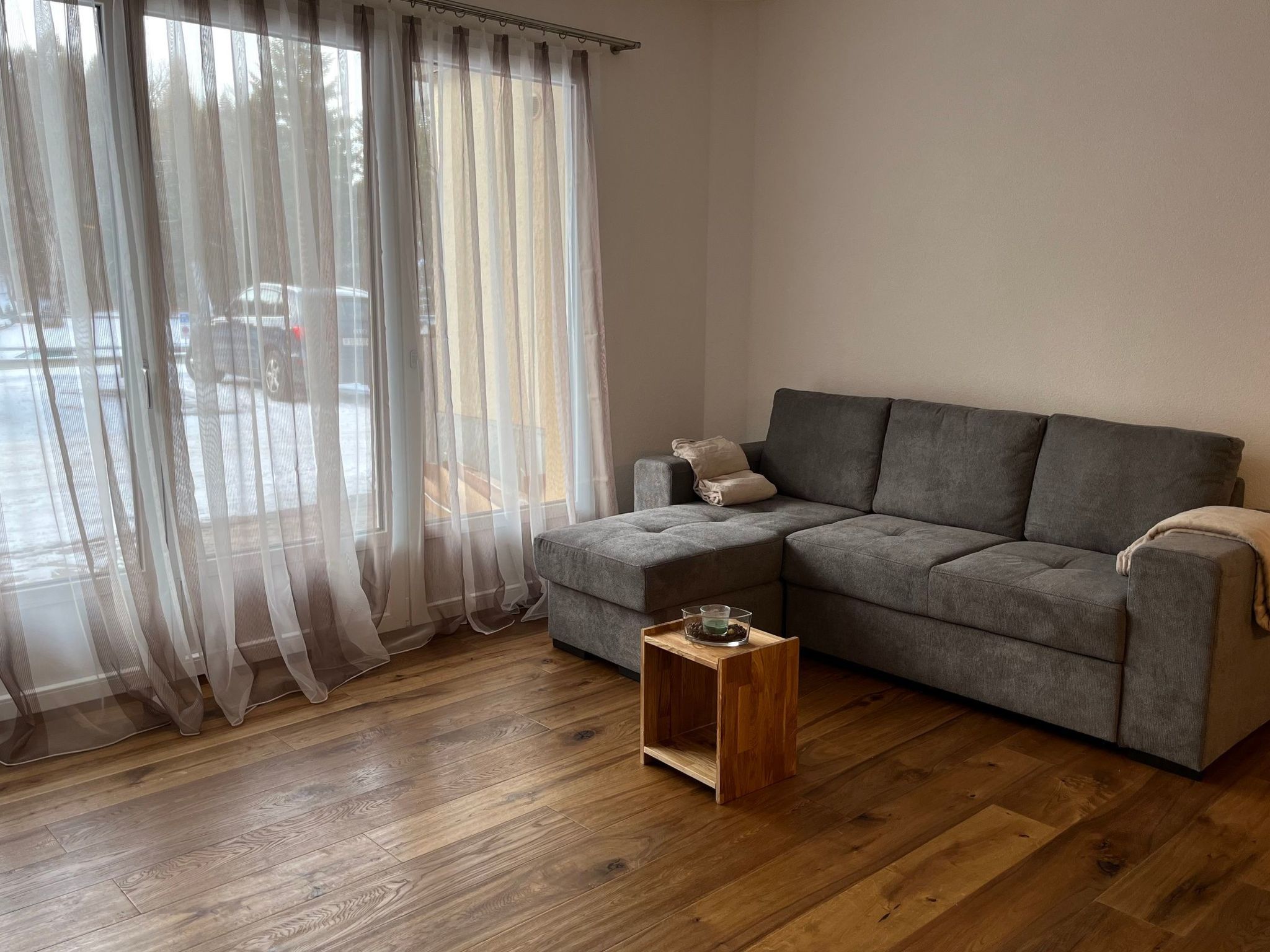 Ein Wohnzimmer mit grauem Sofa, einem kleinen Holztisch, transparenten Vorhängen und Holzboden.