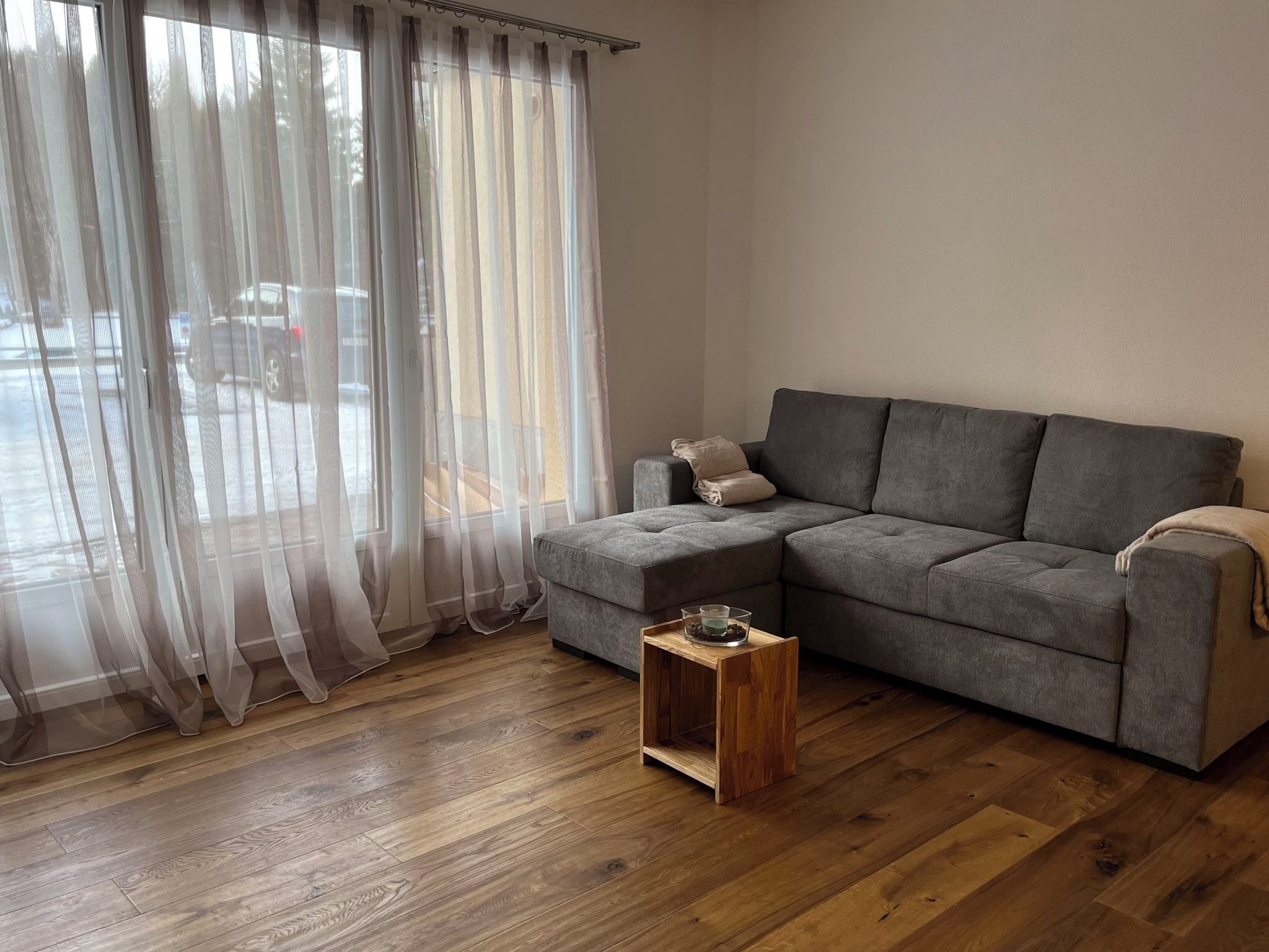 Un salon avec un canapé d’angle gris, une petite table d’appoint en bois, des rideaux légers et un plancher en bois.