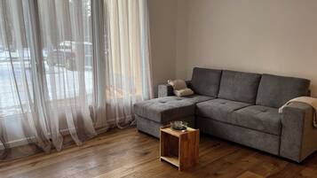 Un salon avec un canapé d’angle gris, une petite table d’appoint en bois, des rideaux légers et un plancher en bois.