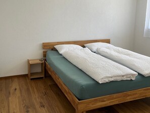 Una cama individual con estructura de madera y colchón verde, con dos almohadas blancas y una manta blanca doblada a los lados.