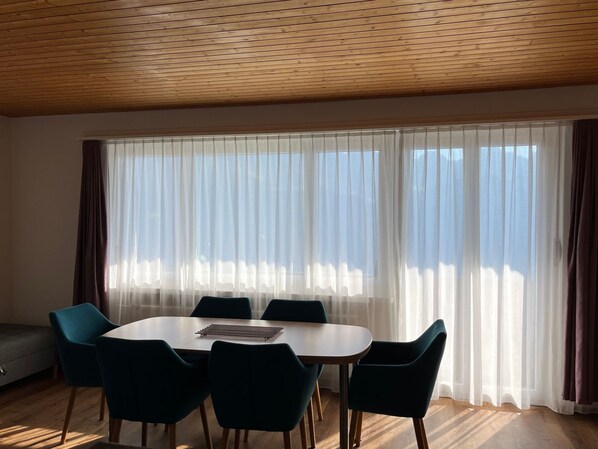 A room with a round table, six chairs, a sofa, and a large window with sheer curtains.