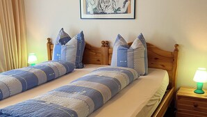 Una cama doble con colcha, dos almohadas, cabecera de madera, mesita de noche con lámpara y un cuadro en la pared.