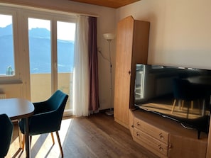 A room with a large flat-screen TV, a wooden cabinet, a dining table with chairs, and a view of mountains through the windows.