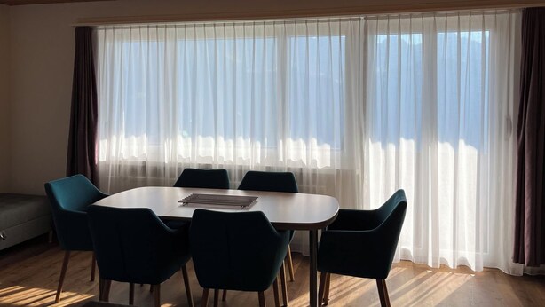 Una habitación con techo de madera, un ventanal con cortinas translúcidas, una mesa redonda con sillas y un sofá.