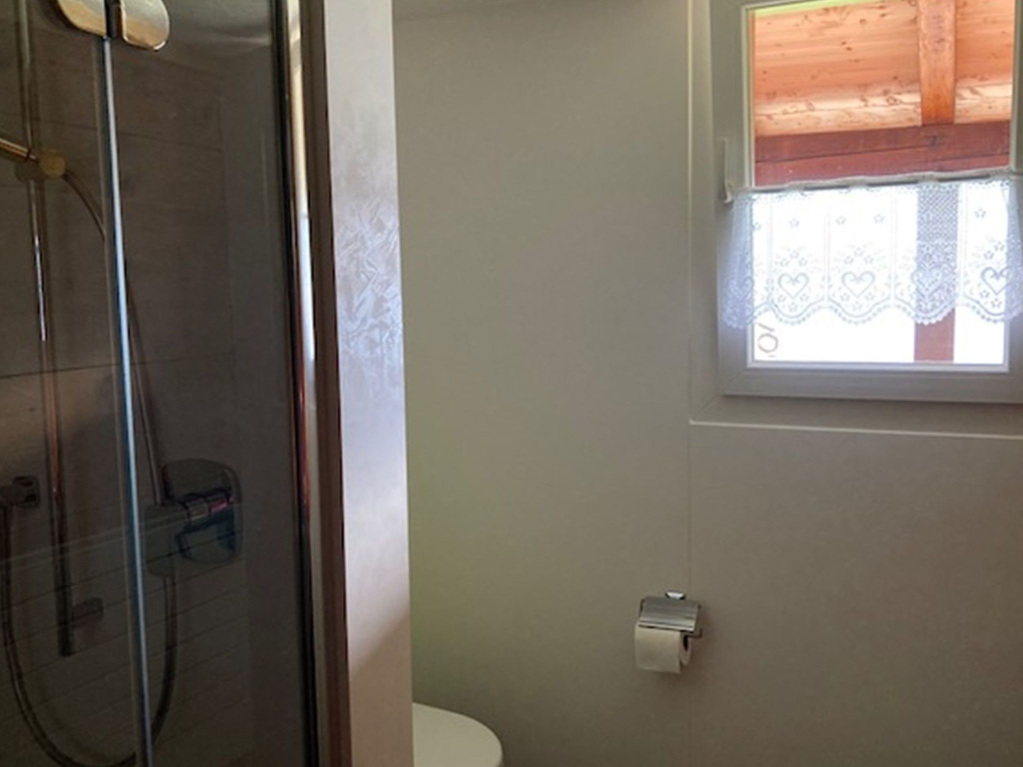 Un baño con cabina de ducha de vidrio, inodoro y ventana con cortinas de encaje.