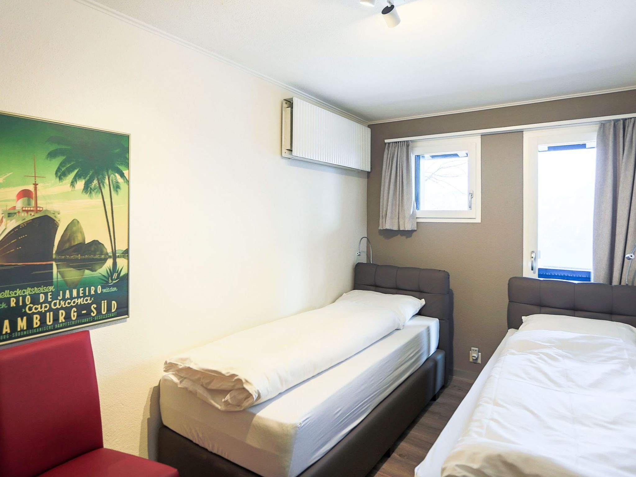 Une chambre d’hôtel avec deux lits simples, une chaise rouge, une affiche au mur et une fenêtre avec des rideaux.