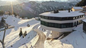 Een besneewd berglandschap met een modern gebouw, grote ramen, een skilift en een helder uitzicht op de omliggende bergen.