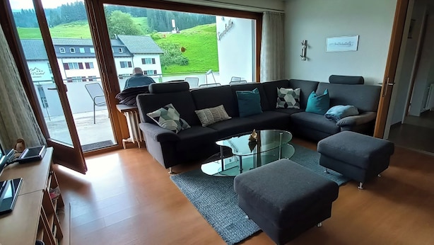 Una sala moderna con un sofá modular, un reposapiés, una mesa de centro de vidrio y vistas a un paisaje verde a través de ventanas amplias.