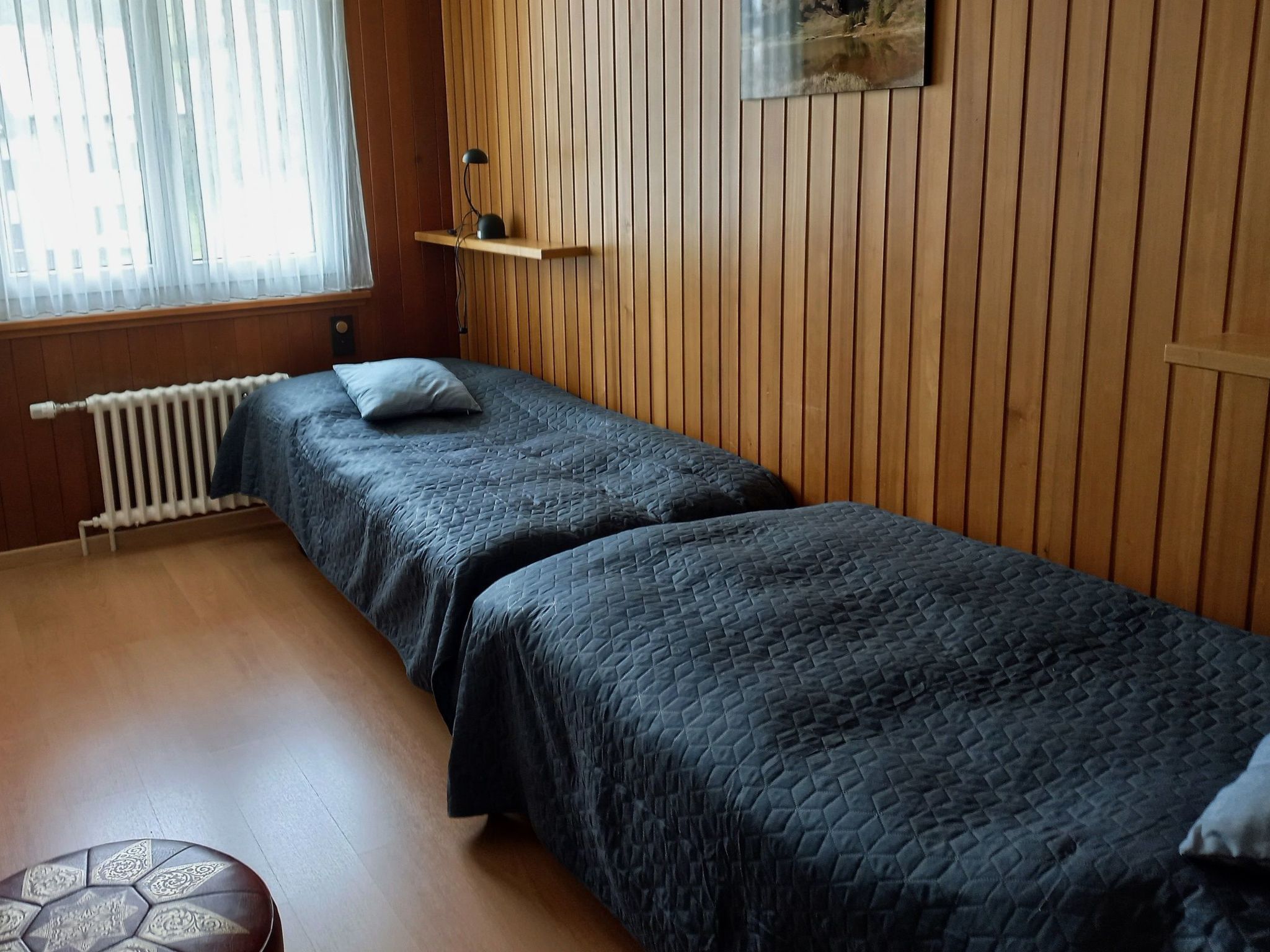 Una habitación con dos camas individuales, un radiador, una pared de madera y una ventana con cortinas traslúcidas.