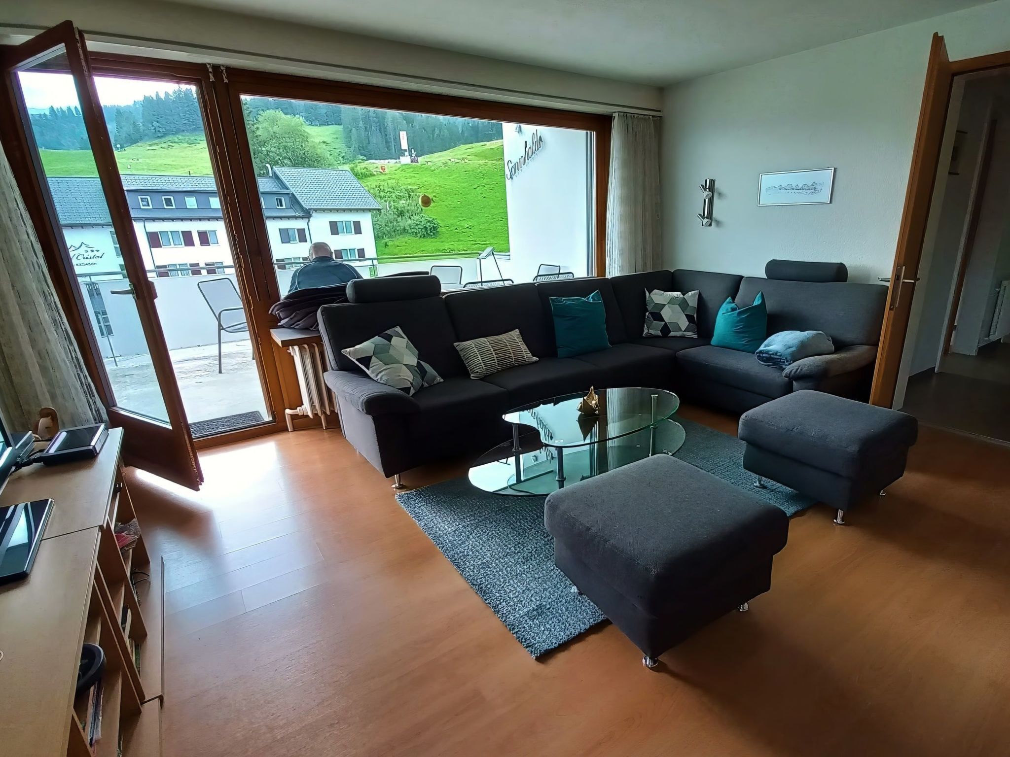 Un salon moderne avec un canapé d’angle, une table basse en verre et une vue sur un paysage verdoyant grâce à de grandes fenêtres.