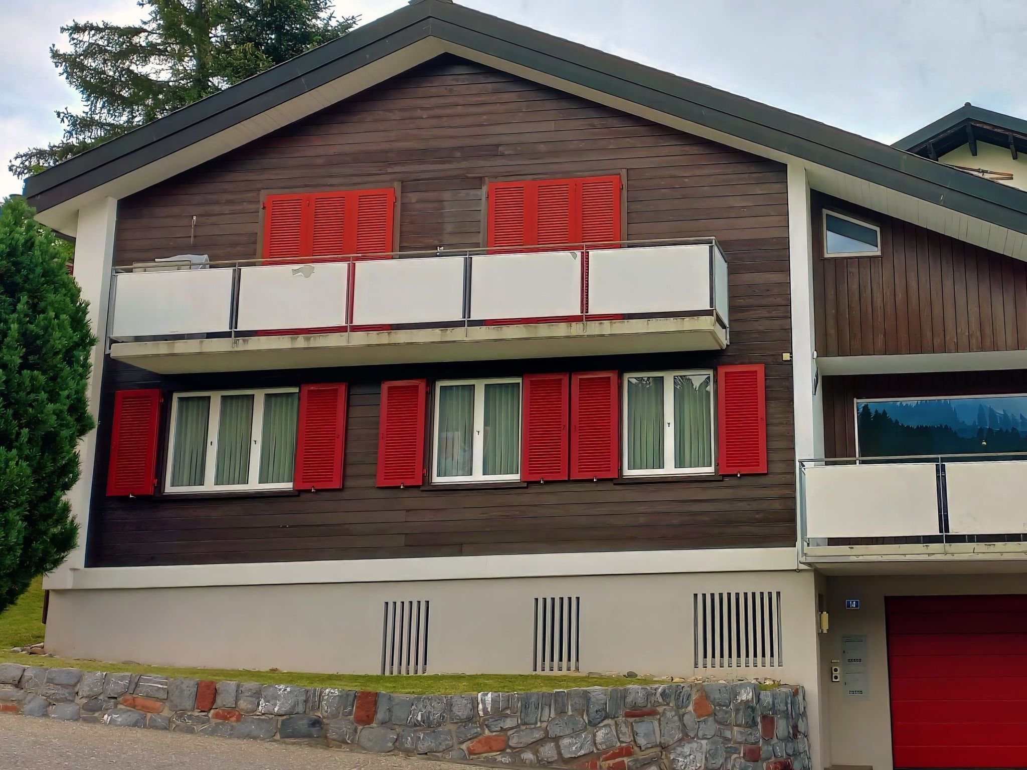 Kaksikerroksinen rakennus, jossa on kivinen pohjarakenne, punaiset ikkunaluukut ja autotalli.