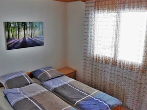 A bedroom with two beds, a nightstand, a painting of a forest, and a window with patterned curtains.