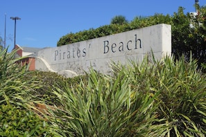 A sign reading "Pirates' Beach" with surrounding greenery and a building in the background. - Seaside Jackalope | Coastal Escape + Large Deck (Galveston)