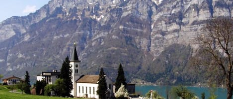 Une église dotée d’un clocher, entourée d’un champ verdoyant et d’un lac, avec des montagnes en arrière-plan.