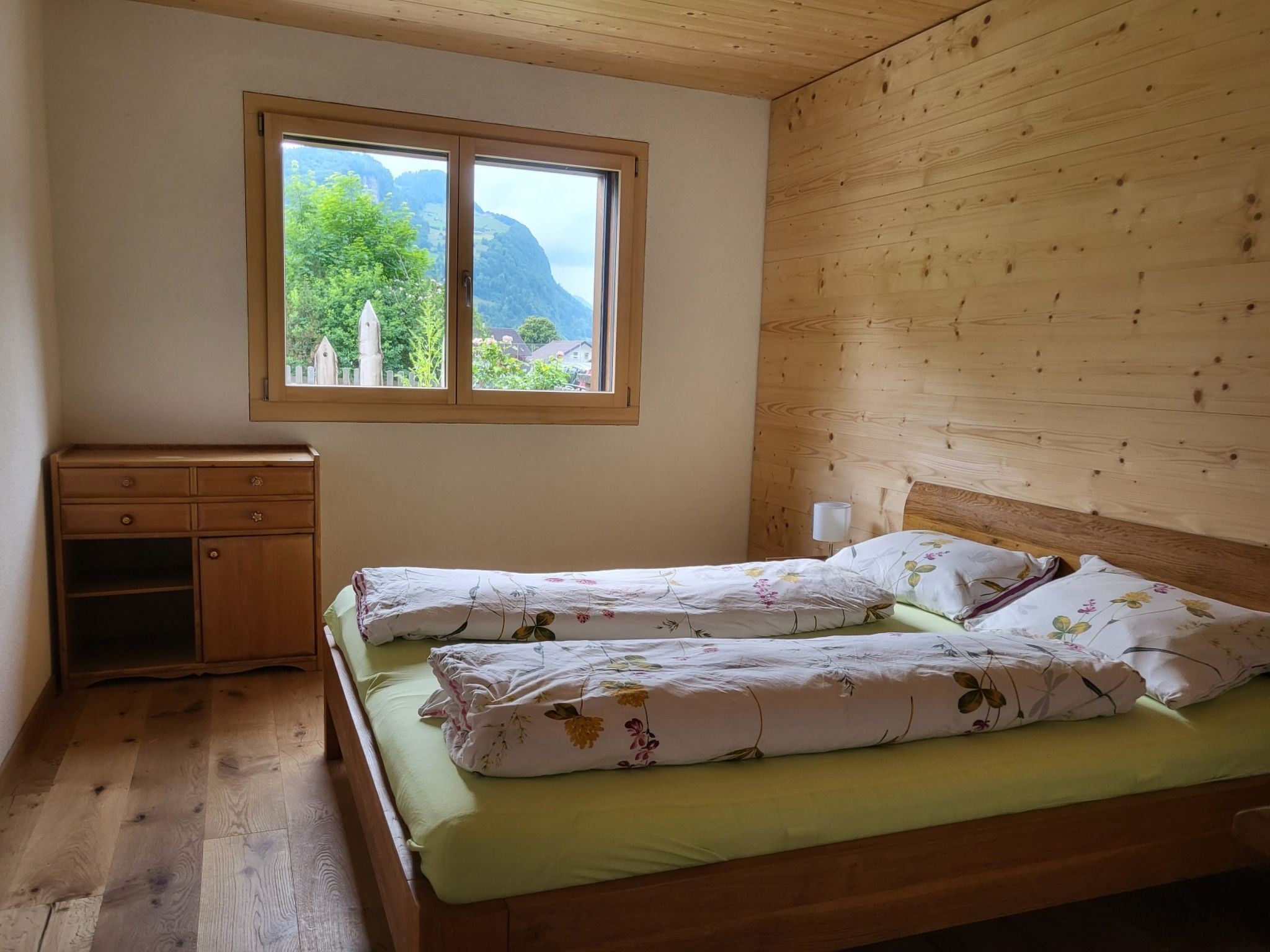 Une chambre à coucher avec une tête de lit en bois, un lit simple avec une literie à motifs floraux, une table de chevet, une fenêtre donnant sur un paysage verdoyant et des montagnes, et une commode en bois.