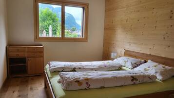 Une chambre à coucher avec une tête de lit en bois, un lit simple avec une literie à motifs floraux, une table de chevet, une fenêtre donnant sur un paysage verdoyant et des montagnes, et une commode en bois.