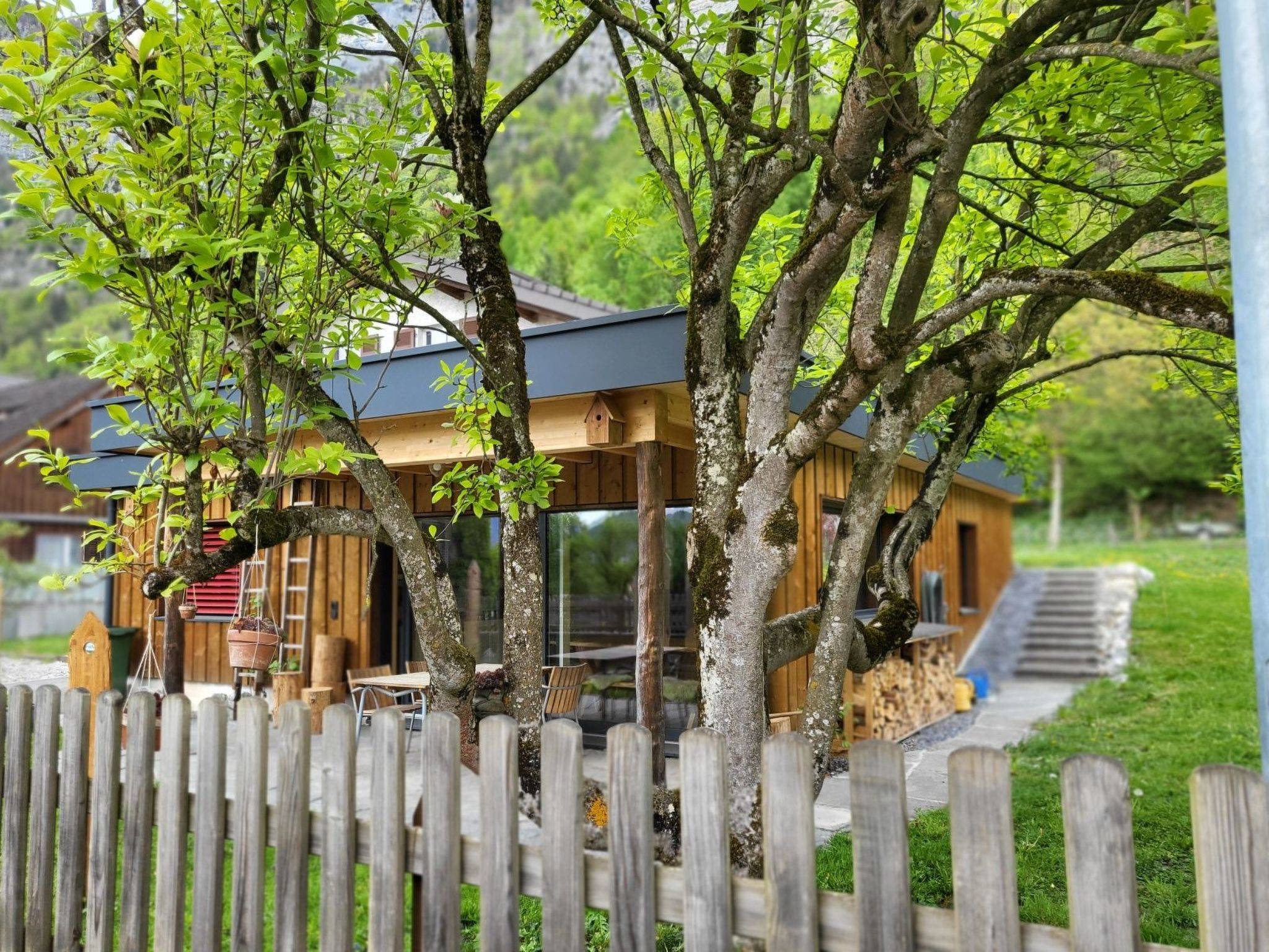 A wooden gazebo with a roof, surrounded by a wooden fence, and a house with a visible staircase in the background.