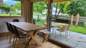 Une table à manger en bois, avec des chaises, de grandes fenêtres et une vue sur un jardin verdoyant et le clocher d’une église.