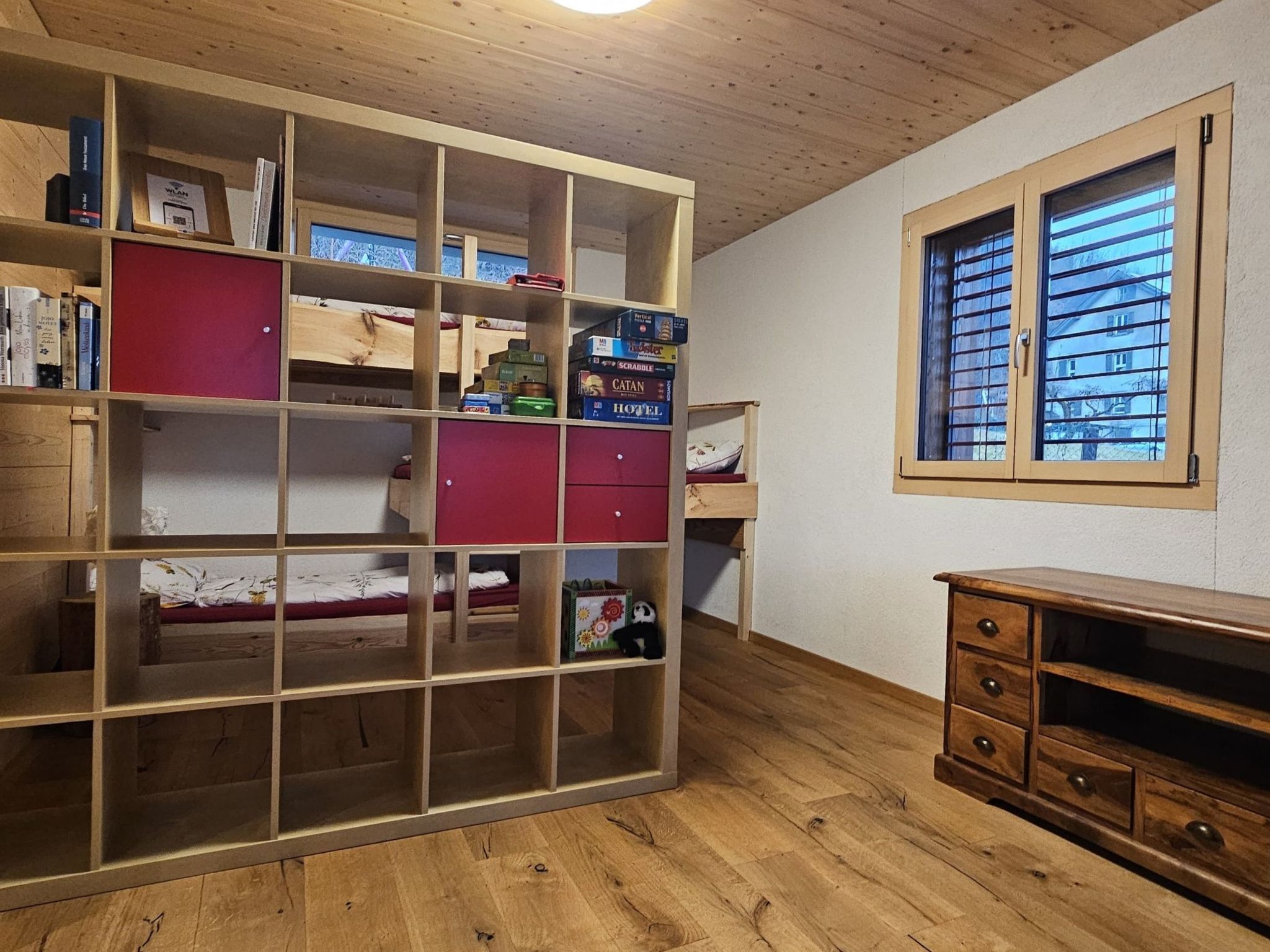 Ein Zimmer mit einem großen Holzregal, einem Fenster mit Jalousien und einer Holzkommode.