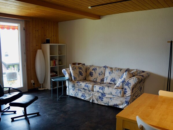 A room with a wooden ceiling, a sofa with floral upholstery, a small table, and a bookshelf.
